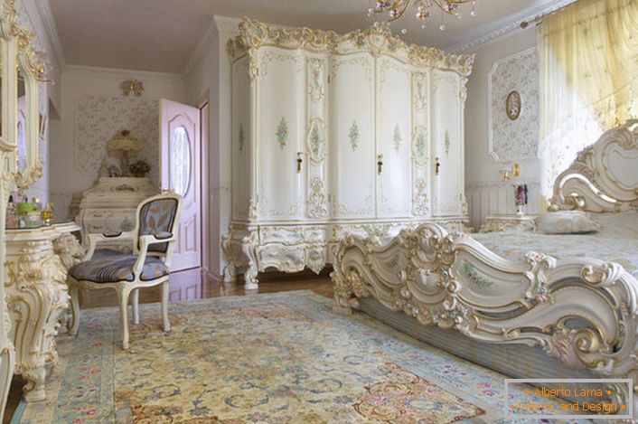 Snežno bela spavaća soba sa rezbarenim masivnim nameštajem od drveta. Krevet sa visokom plafonskom pločom na ploči, elegantno se uklapa u unutrašnjost u baroknom stilu.
